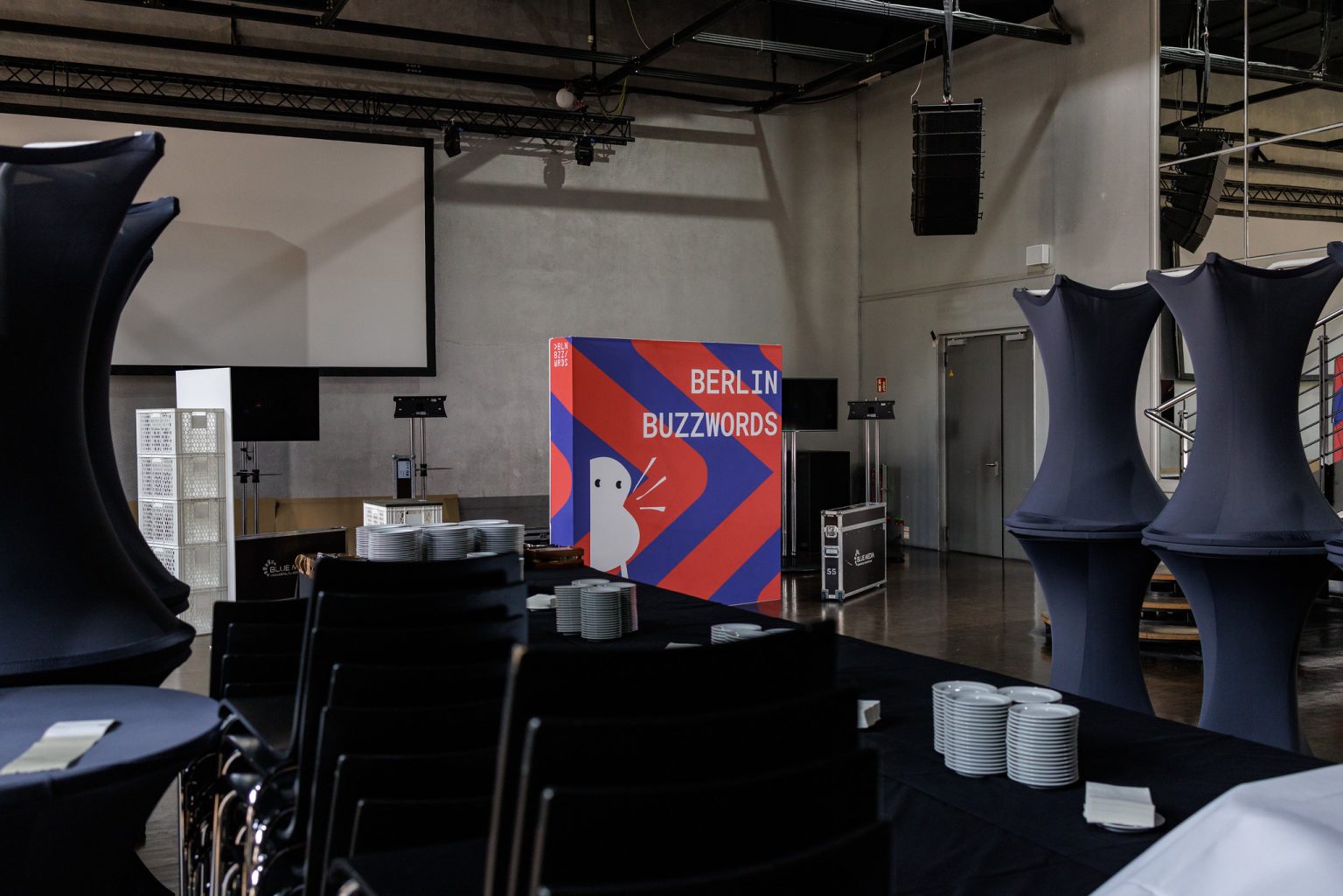 Berlin Buzzwords during build-up: Tables and event equipment is being prepared in the main catering room. In the center there is a colorful Berlin Buzzwords backdrop.